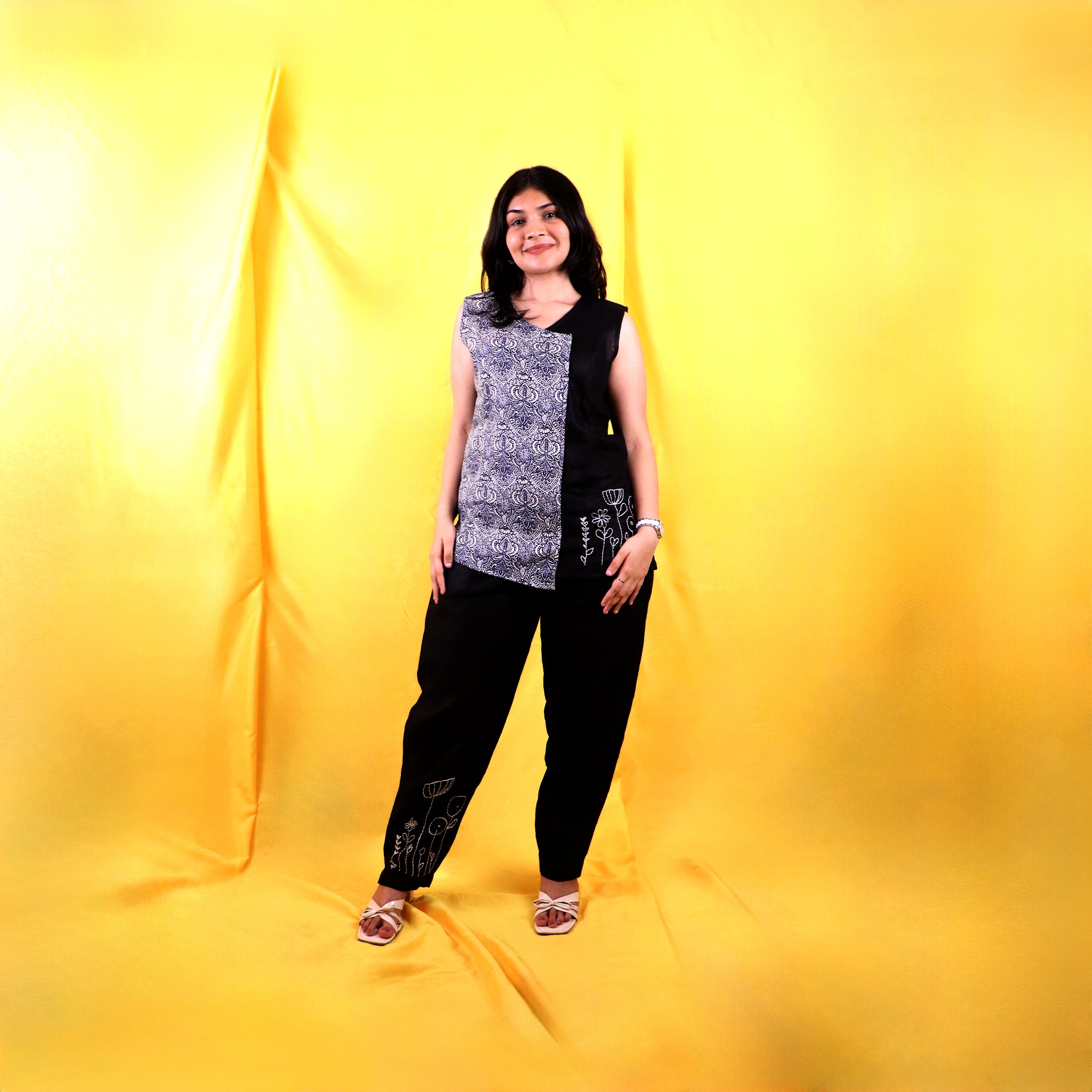 Woman standing against a yellow backdrop wearing a black and white sleeveless linen top with black pants.