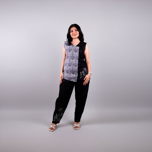 Woman standing against a yellow backdrop wearing a black and white sleeveless linen top with black pants.