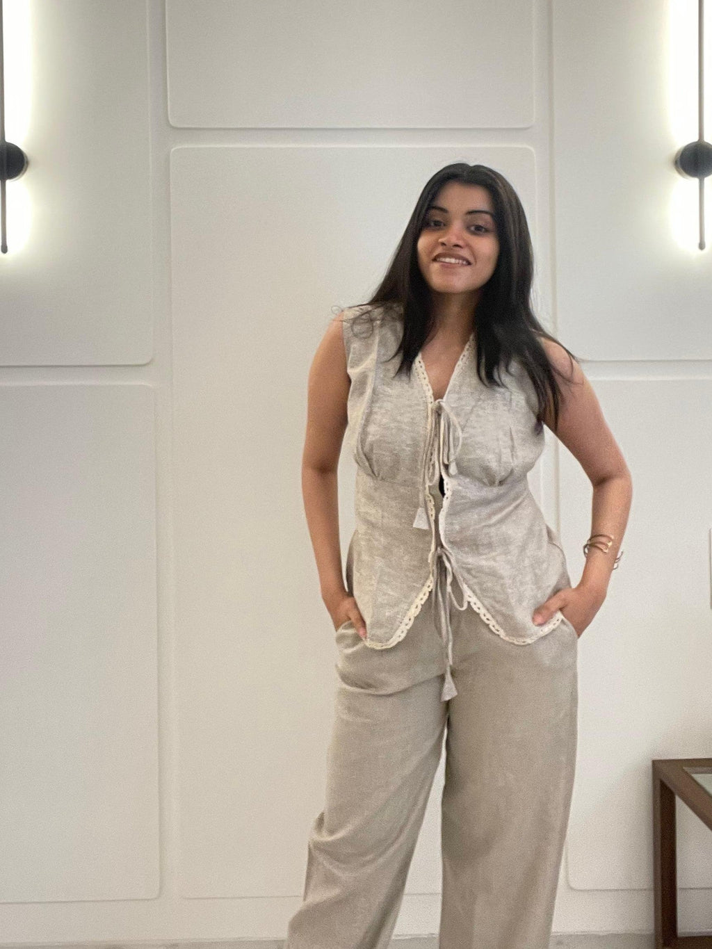A woman wearing a beige linen waistcoat, sleeveless, and pants in a room with white walls.