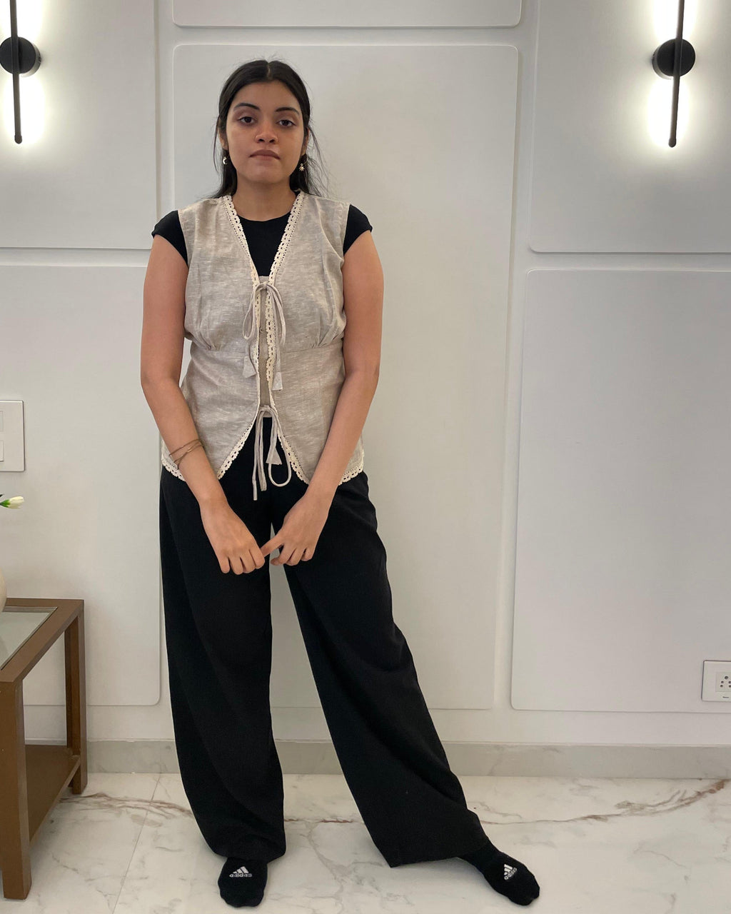 Person wearing a light-colored linen waistcoat over a black top and pants in a room with white walls and a wooden side table.