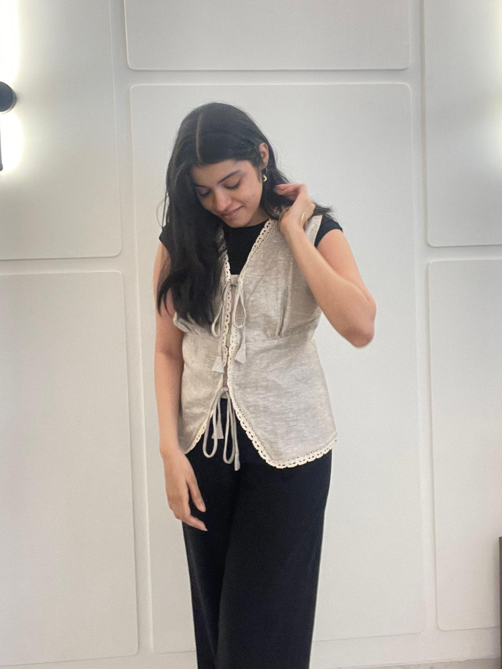 A woman wearing a beige lace linen waistcoat over a black top and pants in front of a white wall.