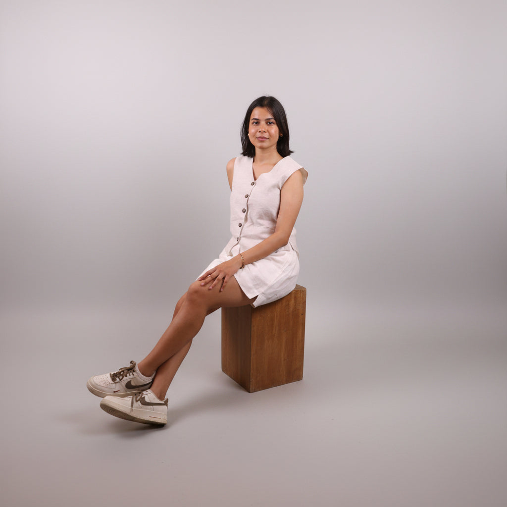 A woman wearing raw linen waistcoat by Sabr India is sitting on a wooden block against a grey background