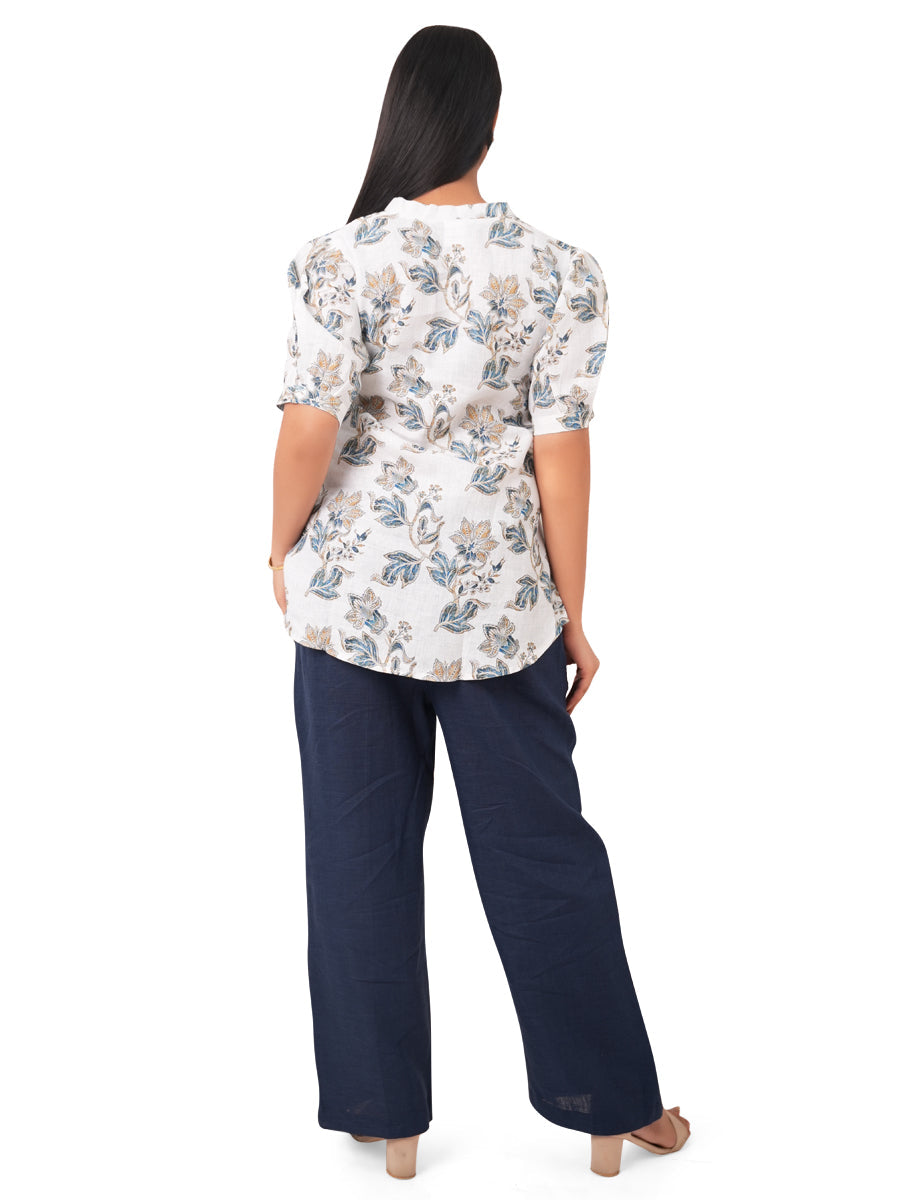 A woman wearing a white floral linen top and navy pants on a white background, back side